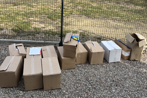 Zabezpieczone paczki papierosów w kartonach stojących na ziemi przy płocie.