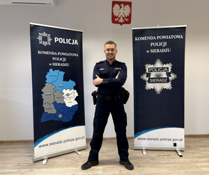 Policjant stoi na tle banerów z logo policji.