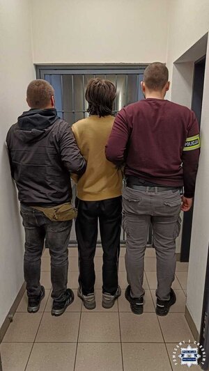 Mężczyzna prowadzony przez policjantów. Widać karaty aresztu.