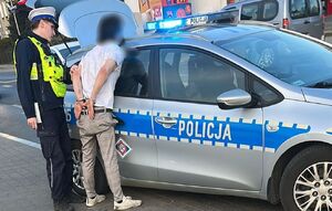 Zatrzymany mężczyzna w kajdankach na rękach stoi z policjantem przy radiowozie.