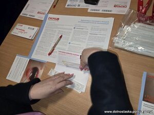 Kobiety siedzą za stołem , pobierany patyczkiem materiał genetyczny. Widać logo policji i fundacji DKMS