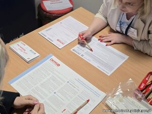 Kobiety siedzą za stołem , pobierany patyczkiem materiał genetyczny. Widać logo policji i fundacji DKMS