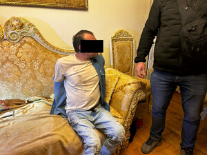 Zatrzymany mężczyzna siedzi na kanapie, obok stoi policjant.