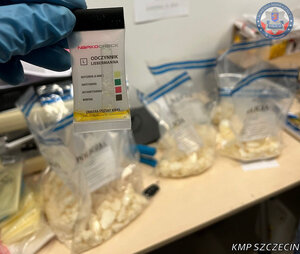 w tle na stole są zabezpieczone narkotyki w foliowych workach. na pierwszym planie widać test narkotykowy, który wskakuje jakiego rodzaju są to narkotyki.