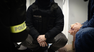 Zatrzymany siedzi na krześle, obok widać policjanta w opasce z napisem Policja. Z prawej strony widać dłonie policjanta.