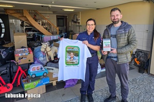 Policjantka stoi z mężczyzną i trzyma koszulkę z wizerunkiem 4-letniej Laury. Z tyłu widać zebrane dary.