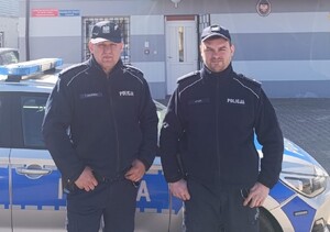 dwóch policjantów pozuje do zdjęcia stoją na tle radiowozu.