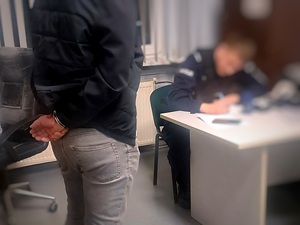 Zatrzymany w kajdankach stoi, przy biurku umundurowany policjant sporządza dokumentację.