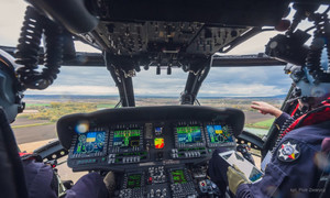 Piloci w kabinie lecącego policyjnego śmigłowca Black Hawk, na środku konsola z ekranami komputerowymi wskazującymi parametry lotu.