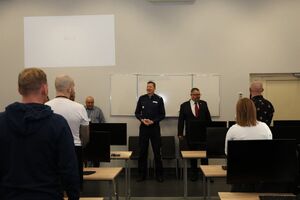 Policjant i mężczyzna w garniturze stoją przodem do zebranych na sali.