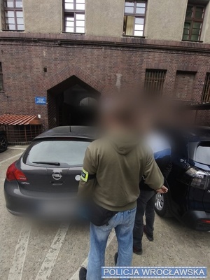 Policjant z zatrzymanym stoi przy samochodzie.