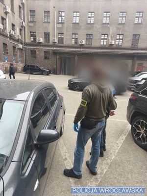 Policjant z zatrzymanym stoi przy samochodzie.