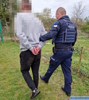 Policjant prowadzi zatrzymanego mężczyznę zakutego w kajdanki.