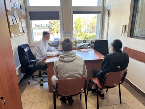 Dwie zatrzymane osoby siedzą na krzesłach w pokoju, w tle widać siedzącego przy biurku policjanta.