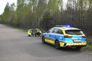 Dwaj policjanci kucają przy stojącej na jezdni hulajnodze elektrycznej, przed nimi widać policyjny radiowóz.