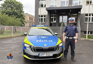 Policjant stoi przy radiowozie. W tle budynek komendy Policji.