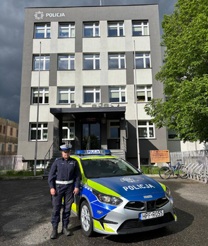 Policjantka stoi przy radiowozie. W tle budynek komendy Policji.