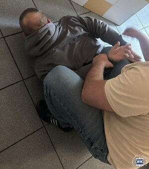 Zatrzymany lezy na ziemi, kuca nad nim policjant, który zakłada mu kajdanki.