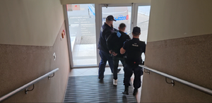 Dwaj policjanci prowadzą zatrzymaną osobę do wyjścia z budynku.