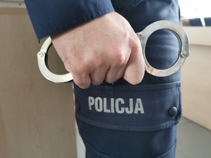 Widok na nogi policjanta i dłoń, w której trzyma kajdanki.
