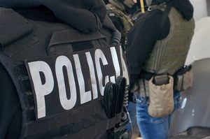Plecy dwóch policjantów, jedne z nich ma na sobie kamizelkę z napisem: Policja.