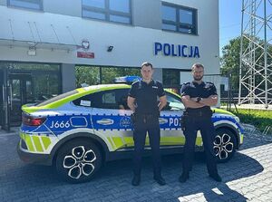 Dwaj umundurowani policjanci pozują do zdjęcia przy radiowozie.