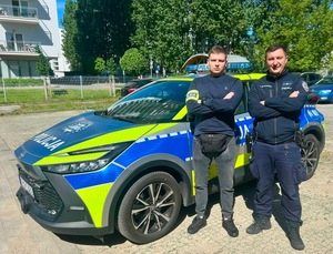 Dwaj policjanci przy radiowozie pozują do zdjęcia.