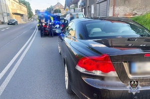 Na pierwszym planie auto na jezdni, a przed nim inne auto, przy którego otwartym bagażniku stoją policjanci.