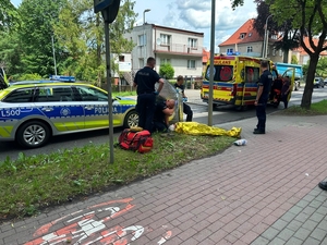Poszkodowany w wypadku leży na trawniku przy ulicy przykryty kocem ratunkowym. Przy nim stoją policjanci. Na ulicy obok stoi radiowóz policyjny i karetka pogotowia.