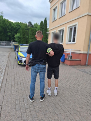 Policjant prowadzi zatrzymanego mężczyznę zakutego w kajdanki.