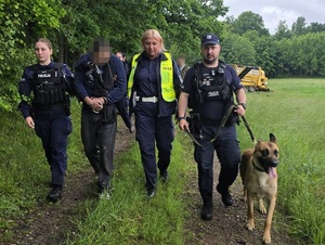 Dwie policjantki prowadzą zatrzymanego. Obok idzie policjant z psem policyjnym.