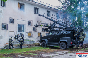 przed białym budynkiem stoi czarny pojazd tur, a na nim są policjanci.