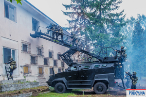 przed białym budynkiem stoi czarny pojazd tur, a na nim są policjanci.