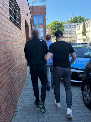 Policjant prowadzi zatrzymanego. Z boku widoczny  jest zaparkowany wzdłuż chodnika policyjny radiowóz.