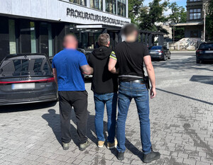 Policjanci prowadzą zatrzymaną osobę, która na rękach trzymanych z tyłu ma założone kajdanki.