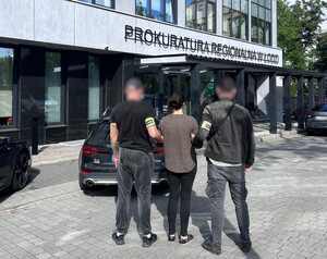 Policjanci prowadzą zatrzymaną osobę, która na rękach trzymanych z tyłu ma założone kajdanki.