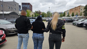 Policjanci prowadzą zatrzymaną osobę, która na rękach trzymanych z tyłu ma założone kajdanki.