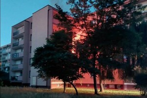 Pomiędzy drzewami prześwituje ogień z płonącego mieszkania w bloku.
