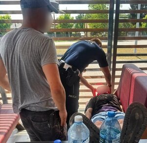 Policjant sprawdza stan mężczyzny leżącego na ławce. Za nim stoi mężczyzna.