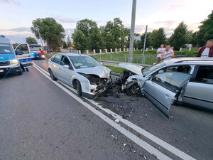 Dwa rozbite auta na drodze po zderzeniu. Na jezdni widać również policyjny radiowóz. Na poboczu stoją ludzie.