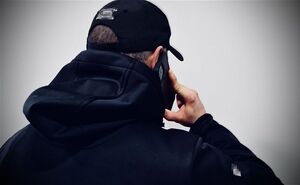 Odwrócony tyłem mężczyzna w kurtce z kapturem. Na głowie ma czapkę typu bejsbolówka. Przy uchu trzyma telefon komórkowy.