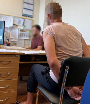 Zatrzymany mężczyzna w kajdankach siedzi na krześle przy biurku policjanta, który go przesłuchuje.