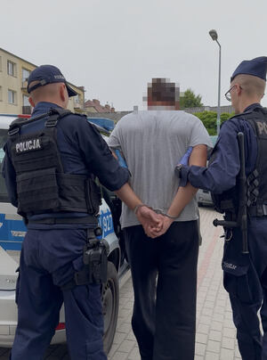 Zatrzymany mężczyzna w kajdankach prowadzony przez dwóch policjantów.