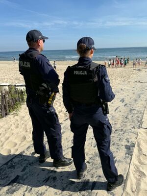 Policjantka i policjant stoją na plazy i przyglądają się dzieciom, które się bawią nad morzem.