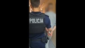 Policjant prowadzi mężczyznę, który na rękach ma kajdanki.