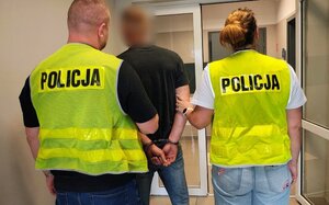 Policjant i policjantka trzymają zatrzymanego mężczyznę zakutego w kajdanki.