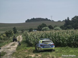 Policyjny radiowóz na polu. Z boku widać jadących na quadzie.