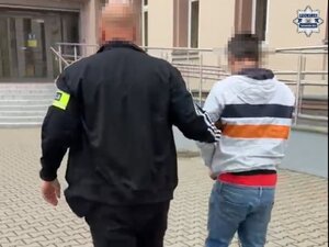 Policjant prowadzi w kierunku budynku zatrzymanego mężczyznę zakutego w kajdanki.