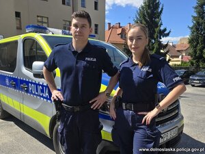 Policjant i policjantka pozują do zdjęcia na tle radiowozu.