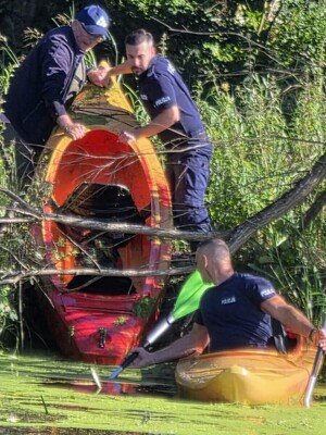 Policjant w kajaku na wodzie, drugi funkcjonariusz stoi na brzegu i wraz z mężczyzną wyciąga z wody drugi kajak.